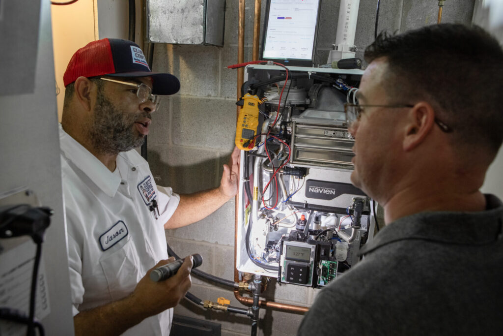 Two men are next to a gas furnace, engaged in a discussion about its functionality and maintenance