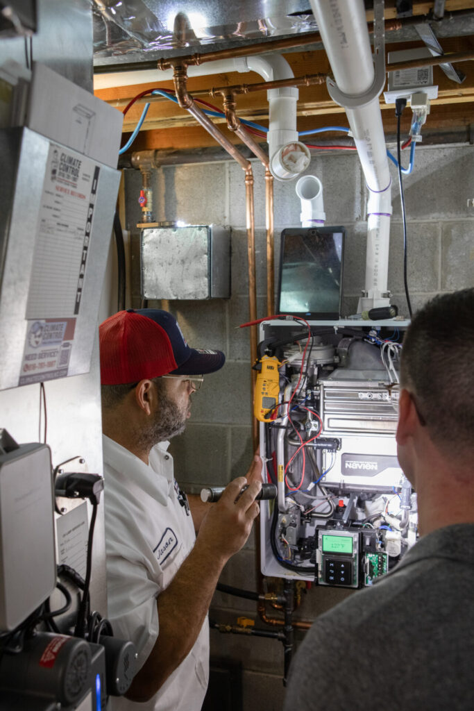 Two men are next to a gas furnace, reviewing its settings and discussing proper usage